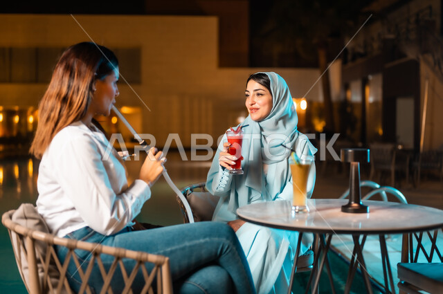 تدخين الشيشة وتناول المشروبات الباردة، اجتماع الصديقات والقيام بنشاطات ممتعة بعطلة نهاية الأسبوع، جلسة وناسة وتسلية في أجواء صيفية لطيفة، صديقتان عربيتان خليجيتان سعوديتان تجلسان أمام المسبح في الهواء الطلق، ارتياد المطاعم والمقاهي في المملكة العربية السعودية