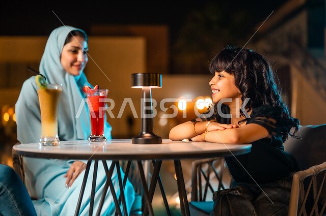 القيام بنشاطات ممتعة بعطلة نهاية الأسبوع، جلسة وناسة وتسلية، أجواء صيفية لطيفة في المطاعم والمقاهي بالمملكة العربية السعودية،أم عربية خليجية سعودية محجبة ترتدي العباءة الملونة برفقة ابنتها الصغيرة تجلسان في المطعم وتتناولان العصائر الباردة المنعشة