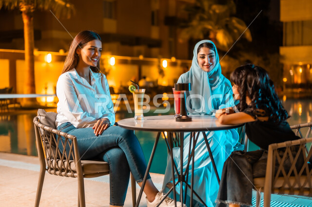 أجواء صيفية لطيفة في المطاعم والمقاهي بالمملكة العربية السعودية، اجتماع الصديقات والقيام بنشاطات ممتعة بعطلة نهاية الأسبوع، جلسة وناسة وتسلية، امرأة عربية خليجية سعودية محجبة ترتدي العباءة الملونة برفقة ابنتها الصغيرة وصديقتها يجلسن في المطعم بالهواء الطلق