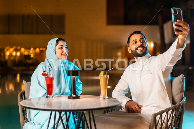 تقوية العلاقات الزوجية وروابط الحب والمودة، التقاط صور السيلفي التذكارية باستخدام الهاتف المحمول، الخروج بسهرة مسائية عائلية، زوجان عربيان خليجيان سعوديان يجلسان أمام المسبح في أحد المنتجعات الفاخرة في السعودية، تناول المشروبات الباردة والعصائر الموسمية اللذيذة