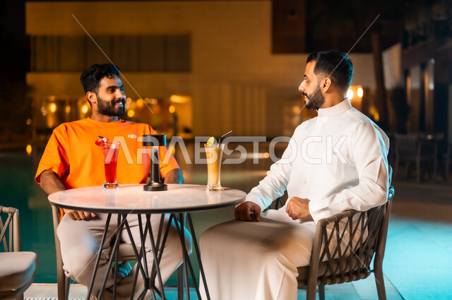 النقاش بين الأصحاب وتبادل أطراف الحديث، المطاعم والكافيهات في المملكة العربية السعودية، قضاء وقت ممتع ومسلي مع الأصدقاء في الخارج، شابان عربيان خليجيان سعوديان يجلسان حول حوض المسبح في مطعم بديكور عصري، الجلسة الخارجية المسائية الهادئة