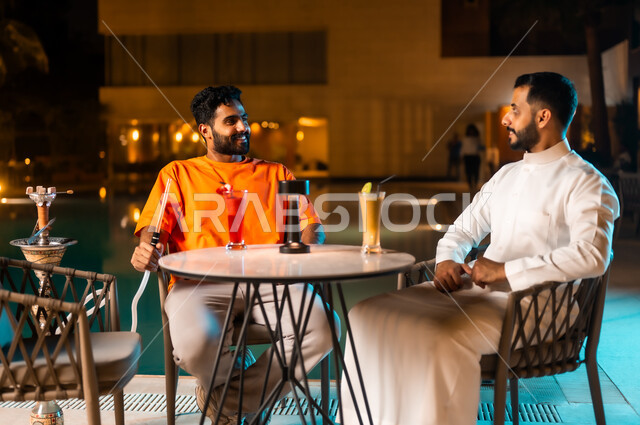 تبادل أطراف الحديث والنقاش، قضاء وقت ممتع ومسلي مع الأصدقاء في الخارج، الكافيهات والمطاعم في المملكة العربية السعودية، شابان عربيان خليجيان سعوديان يجلسان حول حوض المسبح ويدخنان الشيشة في جو شبابي ترفيهي، الاستمتاع بتناول المشروبات الموسمية المنعشة
