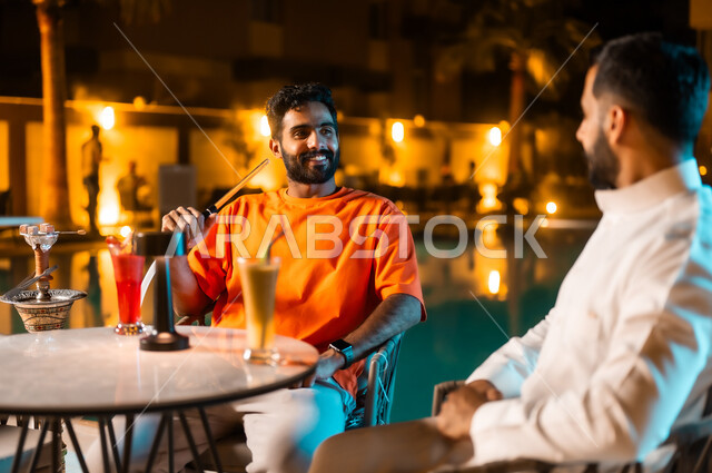 قضاء وقت ممتع ومسلي مع الأصدقاء في الخارج، الكافيهات والمطاعم في المملكة العربية السعودية، شابان عربيان خليجيان سعوديان يجلسان حول حوض المسبح ويدخنان الشيشة في جو شبابي ترفيهي، الاستمتاع بتناول المشروبات الموسمية المنعشة، تبادل أطراف الحديث والنقاش