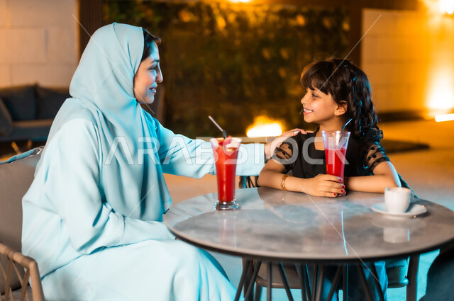 تقوية روابط المودة والحب بين الأم وابنتها، القيام بنشاطات ممتعة بعطلة نهاية الأسبوع، أجواء صيفية لطيفة في المطاعم والمقاهي بالمملكة العربية السعودية،امرأة عربية خليجية سعودية محجبة ترتدي العباءة برفقة طفلتها تجلسان في المطعم وتتناولان العصائر الباردة المنعشة