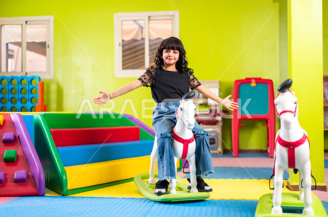 النظر إلى الكاميرا بإيماءات السعادة والسرور، اللعب واللهو والمرح، القيام بنشاط ترفيهي في عطلة نهاية الأسبوع، ارتياد الصالة المغلقة المخصصة للأطفال، فتاة صغيرة عربية خليجية سعودية ترتدي الزي الكاجوال تجلس على الحصان البلاستيكي وتتأرجح وتفتح يديها بحماس