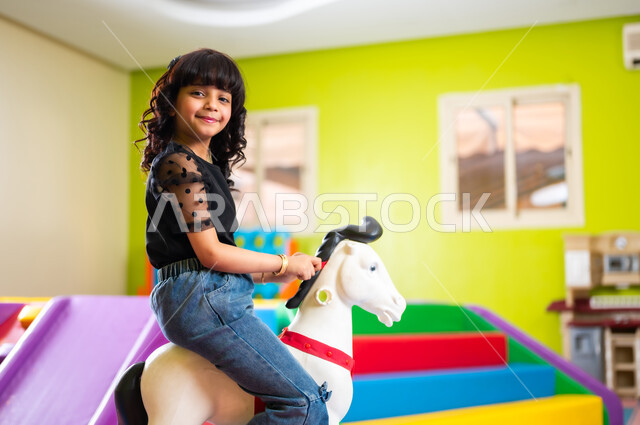 اللعب واللهو والمرح بحماس ونشاط، القيام بنشاطات ترفيهية في عطلة نهاية الأسبوع، ارتياد الصالات المغلقة المخصصة للأطفال، فتاة صغيرة عربية خليجية سعودية ترتدي الزي الكاجوال تجلس على الحصان البلاستيكي وتتأرجح، النظر إلى الكاميرا بإيماءات السعادة والسرور