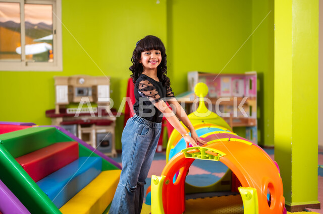 تفريغ الطاقة باللعب واللهو والمرح، القيام بنشاط ترفيهي في عطلة نهاية الأسبوع، ارتياد الصالات المغلقة المخصصة للأطفال، فتاة صغيرة عربية خليجية سعودية ترتدي الزي الكاجوال تلهو وتلعب بحماس ونشاط، النظر إلى الكاميرا بإيماءات السعادة والسرور