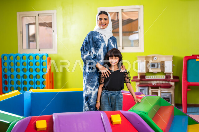 النظر إلى الكاميرا بإيماءات السعادة والسرور، تقوية روابط الأمومة، اصطحاب الأولاد إلى الصالة المخصصة للعب، مشاركة الأبناء لحظات المرح والمتعة، القيام بنشاطات عائلية في عطلة نهاية الأسبوع، فتاة عربية خليجية سعودية ترتدي الزي الكاجوال برفقة والدتها تلعبان سويا