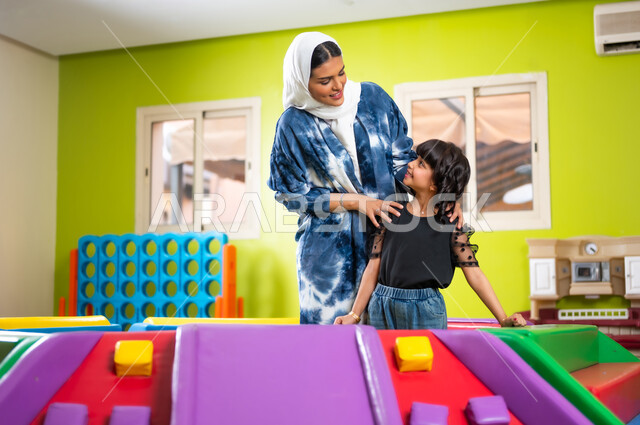 تقوية روابط الأمومة، النظر إلى الكاميرا بإيماءات السعادة والسرور، اصطحاب الأولاد إلى الصالة المخصصة للعب، مشاركة الأبناء لحظات المرح والمتعة، القيام بنشاط عائلي في عطلة نهاية الأسبوع، فتاة عربية خليجية سعودية ترتدي الزي الكاجوال برفقة والدتها تلعبان سويا