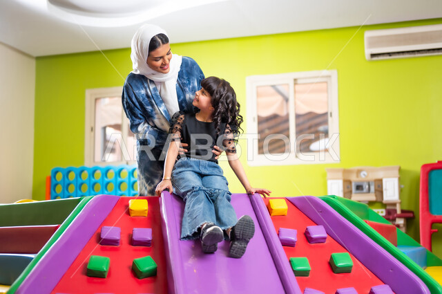 المرح واللعب مع الأبناء ومشاركة اللحظات السعيدة، تعزيز رابط الأمومة بين الأم وابنتها، اصطحاب الأولاد إلى الصالة المخصصة للألعاب، القيام بنشاط عائلي في عطلة نهاية الأسبوع، فتاة عربية خليجية سعودية ترتدي الزي الكاجوال تتزحلق على الزحليقة بمساعدة والدتها