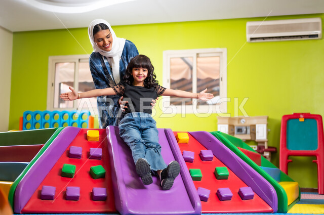 الاستمتاع بمشاركة الأبناء المرح واللعب، تعزيز رابط الأمومة بين الأم وابنتها، اصطحاب الأولاد إلى الصالة المخصصة للألعاب، القيام بنشاط عائلي في عطلة نهاية الأسبوع، فتاة عربية خليجية سعودية ترتدي الزي الكاجوال تتزحلق على الزحليقة بمساعدة والدتها بإيماءات السعادة