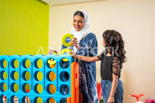 Taking children to children's playrooms, spending fun recreational times, a Saudi Arabian Gulf girl wearing casual clothes with her mother playing together, developing children's mental abilities through intellectual and sensory games, doing family activities on the weekend