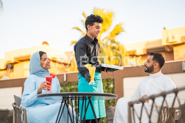 العمل في المطاعم والكافيهات الفاخرة في السعودية، تناول العصائر الموسمية الباردة والمنعشة، نادل يرتدي زي اليونيفورم يقوم بتقديم الطلبات للزبائن، زوجان عربيان خليجيان سعوديان يستمتعان بوقتهما في جلسة خارجية للمطعم ضمن أجواء صيفية لطيفة
