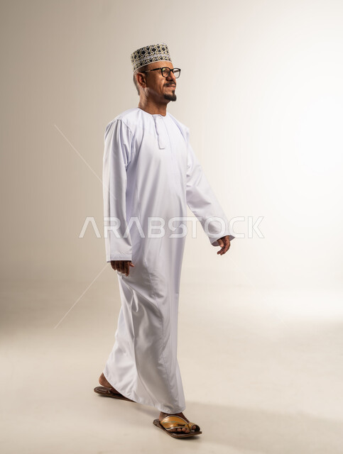 التقدم والتوجه نحو الأمام، الاهتمام بالمظهر الخارجي، ارتداء أزياء عمانية تقليدية، بورتريه من الجانب لرجل عربي خليجي عماني يرتدي الدشداشه و الكمه يلبس النظارات الطبية يمشي وينظر أمامه بإيماءات التركيز, الجسم بالطول الكامل، خلفية بيضاء