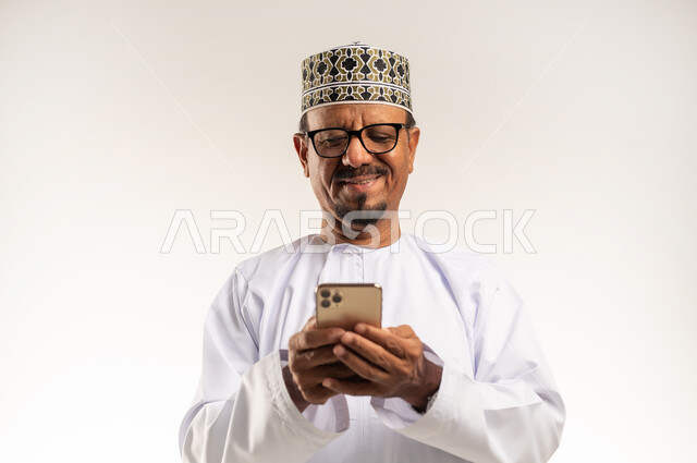 استخدام الأجهزة التقنية والتكنولوجيا الحديثة، التواصل مع الأهل والأصدقاء وسماع اخبارهم، تصفح مواقع التواصل الاجتماعي، بورتريه مقرب لرجل عربي خليجي عماني يرتدي الدشداشة والكمه يحمل بيده الهاتف المحمول بإيماءات السعادة والسرور، خلفية بيضاء