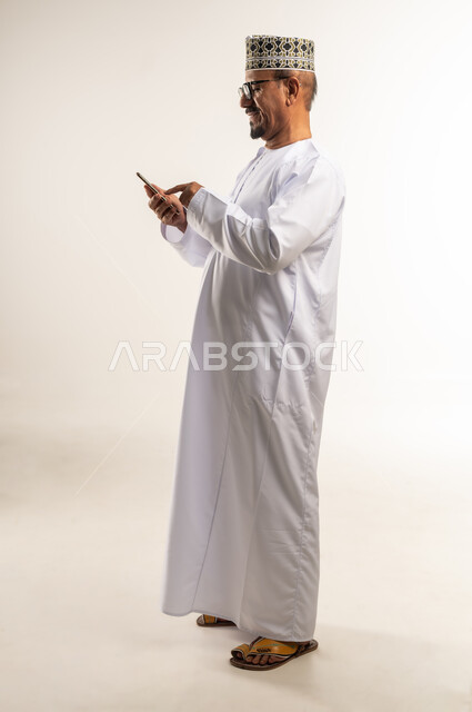 تصفح مواقع التواصل الاجتماعي، التواصل مع الأهل والأصدقاء وسماع اخبارهم، استخدام الأجهزة التقنية الحديثة، بورتريه من الجانب لرجل عربي خليجي عماني يرتدي الدشداشة والكمه يحمل بيده الهاتف المحمول بإيماءات السعادة، الجسم بالطول الكامل، خلفية بيضاء