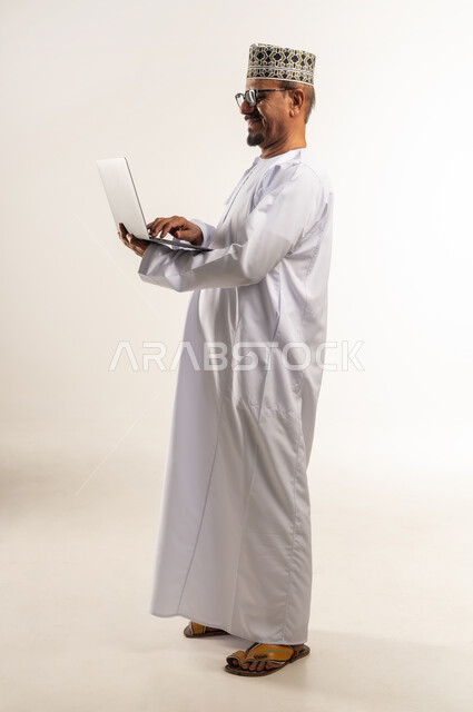 مفهوم العمل عن بعد, إجراء مكالمات الفيديو والاجتماعات أونلاين، استخدام الأجهزة التقنية الحديثة، التواصل مع الأهل والأصدقاء وسماع أخبارهم، بورتريه من الجانب لرجل عربي خليجي عماني يرتدي الدشداشة والكمه يحمل بيده الحاسوب المحمول، الجسم بالطول الكامل, خلفية بيضاء