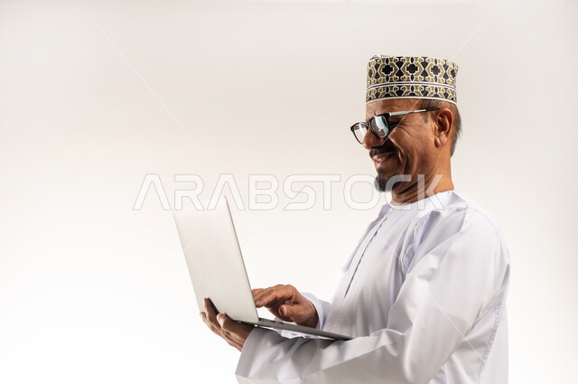 Communicating with family and friends and hearing their news, using modern technical devices, the concept of remote work, making video calls and online meetings, close-up portrait from the side of an Arab Gulf Omani man wearing a dishdasha and a kuma holding a laptop in his hand with gestures of pleasure, white background