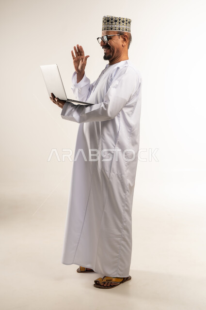 إجراء مكالمة الفيديو والاجتماع أونلاين، استخدام الأجهزة التقنية الحديثة، مفهوم العمل عن بعد, التواصل مع الأهل والأصدقاء وسماع أخبارهم، بورتريه من الجانب لرجل عربي خليجي عماني يرتدي الدشداشة والكمه يحمل بيده الحاسوب المحمول، الجسم بالطول الكامل, خلفية بيضاء