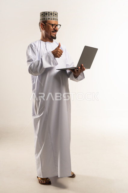 رفع اصبع الإبهام بعلامة الجودة والامتياز، مفهوم العمل عن بعد, استخدام جهاز تقني حديث، إجراء مكالمة فيديو واجتماع أونلاين، بورتريه مقرب من الجانب لرجل عربي خليجي عماني يرتدي الدشداشة والكمه يحمل بيده الحاسوب المحمول، الجسم بالطول الكامل, خلفية بيضاء