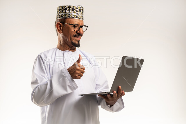 Remote work concept, thumb up sign, using modern technology device, video call and online meeting, close-up side portrait of an Arab Gulf Omani man wearing a dishdasha and a sleeve holding a laptop, white background