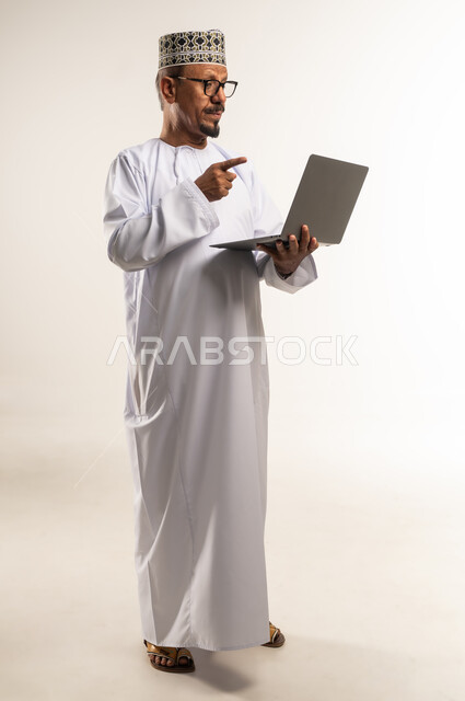 Using modern technology device, remote work concept, pointing index finger at laptop screen, making video call and online meeting, side portrait of an Arab Gulf Omani man wearing a dishdasha and a sleeve holding a laptop in his hand, full body, white background