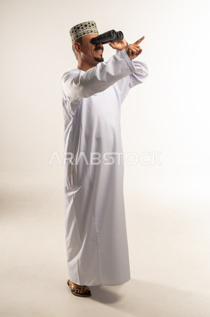 الإشارة باصبع السبابة إلى مكان أو عنصر ما بعيد، توضيح الرؤية وتقريبها عبر التلسكوب, حب الاستكشاف و المغامرة, بورتريه من الجانب لرجل عربي خليجي عماني يرتدي الدشداشة والكمه يحمل بيده منظار ثنائي العينين و ينظر الى شيء ما، الجسم بالطول الكامل, خلفية بيضاء
