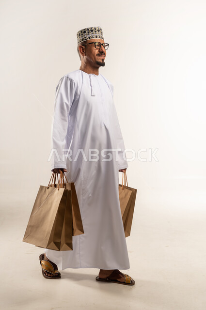 تعابير الثقة و السعادة للاستفادة من العروض و التخفيضات, بورتريه من الجانب لرجل عربي خليجي عماني يرتدي الدشداشة والكمه يحمل بيده الأكياس الورقية ويمشي نحو الأمام, متعة التسوق في مولات ومتاجر السلطنة، اقتناء المستلزمات والهدايا, الجسم بالطول الكامل, خلفيه بيضاء