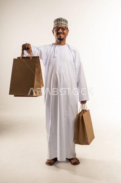 النظر إلى الكاميرا بثقة و سعادة للاستفادة من العروض و التخفيضات, بورتريه لرجل عربي خليجي عماني مبتسم يرتدي الدشداشة والكمه يحمل بيده الأكياس الورقية, متعة التسوق في مولات ومتاجر السلطنة، اقتناء المستلزمات و الهدايا, الجسم بالطول الكامل, خلفيه بيضاء