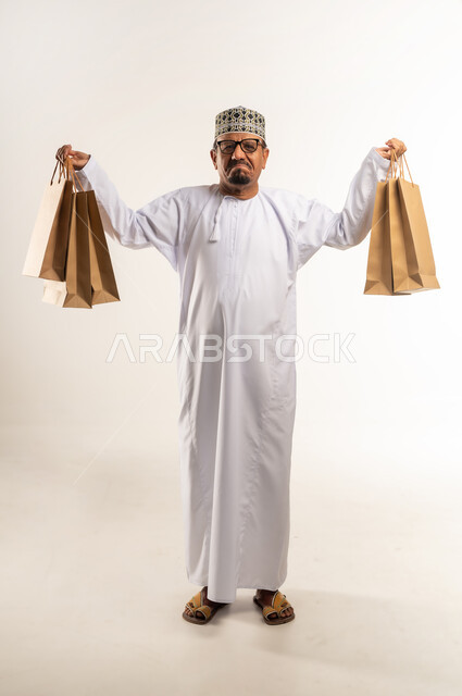 Gestures of confusion and lack of knowledge, shopping in malls and stores of the Sultanate, taking advantage of offers and discounts, portrait of a smiling Arab Gulf Omani man wearing a dishdasha and a kummah holding paper bags in his hand and raising his hands in the air, acquiring supplies and gifts, full body, white background