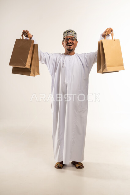 التعبير عن السعادة بالاستفادة من العروض و التخفيضات, بورتريه لرجل عربي خليجي عماني مبتسم يرتدي الدشداشة والكمه يحمل بيده الأكياس الورقية ويرفع يديه في الهواء, متعة التسوق في مولات ومتاجر السلطنة، اقتناء المستلزمات و الهدايا, الجسم بالطول الكامل, خلفيه بيضاء