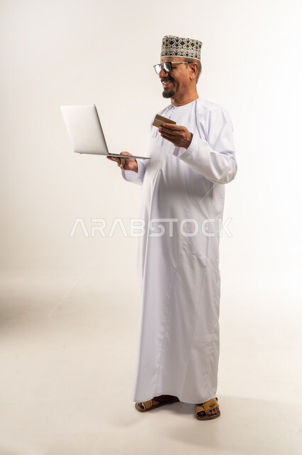 مفهوم التسوق الالكتروني, الحجز و الطلب عبر الانترنت, استخدام الأجهزة التقنية الحديثة بالبيع والشراء، الدفع عن طريق البطاقة البنكية، بورتريه لرجل عربي خليجي عماني يرتدي الدشداشة والكمه يحمل بيده الحاسوب المحمول بإيماءات السرور، الجسم بالطول الكامل، خلفية بيضاء