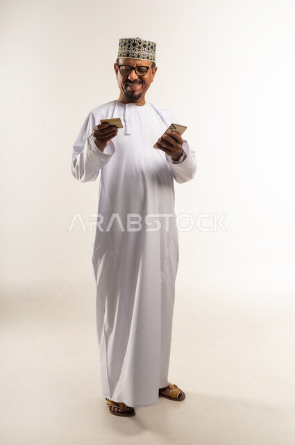 استخدام جهاز تقني حديث بالبيع والشراء، مفهوم التسوق الالكتروني, الحجز و الطلب عبر الانترنت, الدفع عن طريق البطاقة البنكية، بورتريه لرجل عربي خليجي عماني يرتدي الدشداشة والكمه يحمل بيده الهاتف المحمول بإيماءات السرور، الجسم بالطول الكامل، خلفية بيضاء