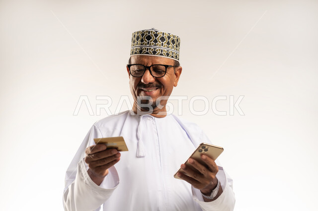 Payment by bank card, use of modern technical devices in buying and selling, concept of e-shopping, booking and ordering via the Internet, close-up portrait of an Arab Gulf Omani man wearing a dishdasha and a kummah holding a mobile phone with gestures of pleasure, white background