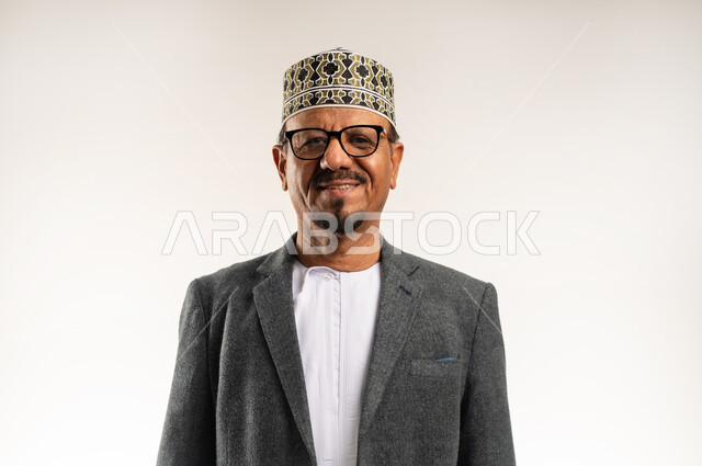 ارتداء ملابس الشتاء الدافئة، الاهتمام بالمظهر الخارجي، الوقوف بتعابير الثقة بالنفس، بورتريه مقرب لرجل عربي خليجي عماني يرتدي الدشداشة والكمه والمعطف الشتوي ينظر إلى الكاميرا بإيماءات السرور والبشاشة، خلفية بيضاء