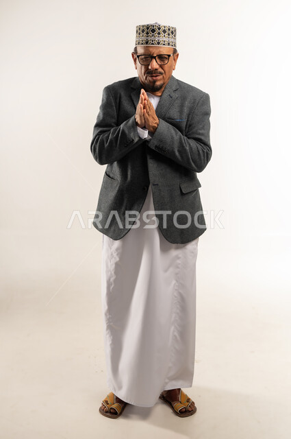 الحاجة لتدفئة اليدين، ارتداء ملابس الشتاء الدافئة، الشعور بالبرد الشديد والصقيع، بورتريه لرجل عربي خليجي عماني يرتدي الدشداشة والكمه والمعطف الشتوي يضم يديه إلى بعضهما في محاولة لرفع الحرارة، الجسم بالطول الكامل، خلفية بيضاء