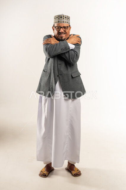 وضع اليدين على الكتفين بشكل متقاطع، ارتداء ملابس الشتاء الدافئة، الشعور بالبرد الشديد والصقيع، بورتريه لرجل عربي خليجي عماني يرتدي الدشداشة والكمه والمعطف الشتوي يضم يديه إلى جسده في محاولة لتدفئة نفسه، الجسم بالطول الكامل، خلفية بيضاء