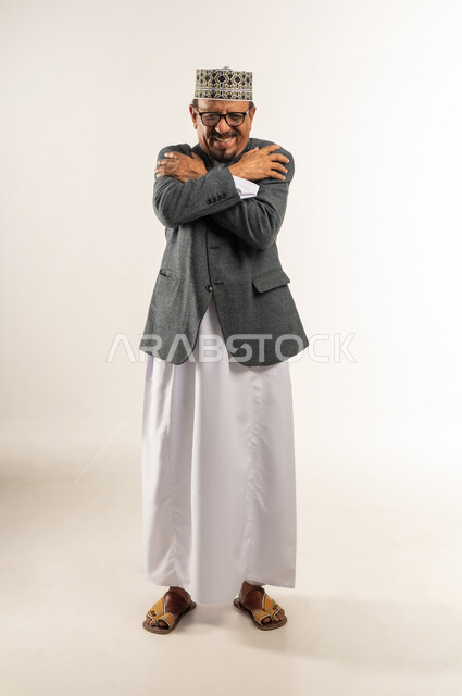 الأجواء الشتوية الباردة والصقيع، وضع اليدين على الكتفين بشكل متقاطع، ارتداء ملابس الشتاء الدافئة، بورتريه لرجل عربي خليجي عماني يرتدي الدشداشة والكمه والمعطف الشتوي يضم يديه إلى جسده في محاولة لتدفئة نفسه، الجسم بالطول الكامل، خلفية بيضاء