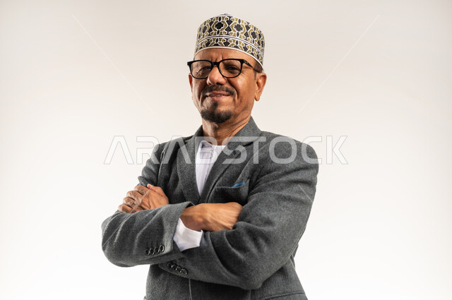 الوقوف بأيدي متقاطعة بتعابير الثقة بالنفس، ارتداء ملابس الشتاء الدافئة، الاهتمام بالمظهر الخارجي، بورتريه مقرب لرجل عربي خليجي عماني يرتدي الدشداشة والكمه والمعطف الشتوي ينظر إلى الكاميرا بإيماءات السرور والبشاشة، خلفية بيضاء