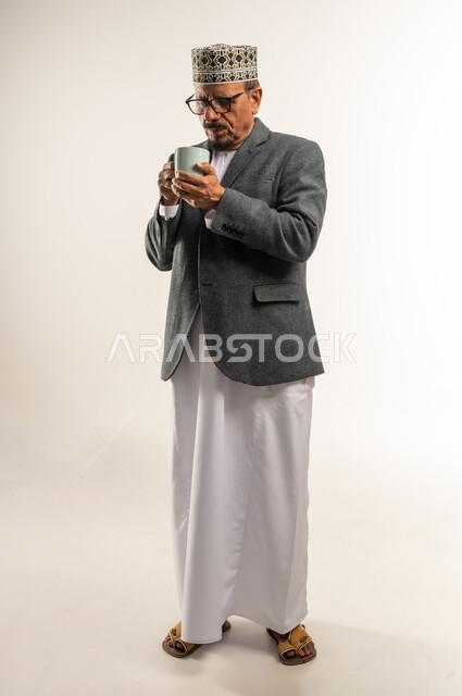 محاولة التغلب على الشعور بالبرد والصقيع، تناول مشروب ساخن بالأجواء الشتوية الباردة، ارتداء ملابس الشتاء الثقيلة، بورتريه لرجل عربي خليجي عماني يرتدي الكمه والمعطف الشتوي يمسك كوب من القهوة الدافئة، الجسم بالطول الكامل، خلفية بيضاء