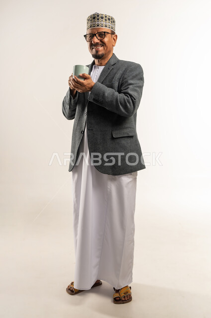 النظر إلى الكاميرا بإيماءات السرور، التغلب على الشعور بالبرد والصقيع، تناول مشروب ساخن بالأجواء الشتوية الباردة، ارتداء ملابس الشتاء الثقيلة، بورتريه لرجل عربي خليجي عماني يرتدي الكمه والمعطف الشتوي يمسك كوب من القهوة الدافئة، الجسم بالطول الكامل، خلفية بيضاء