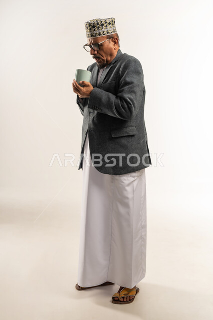 تناول مشروب ساخن بالأجواء الشتوية الباردة، التغلب على الشعور بالبرد والصقيع، ارتداء ملابس الشتاء الثقيلة، بورتريه لرجل عربي خليجي عماني يرتدي الكمه والمعطف الشتوي يمسك كوب من القهوة الدافئة، الجسم بالطول الكامل، خلفية بيضاء