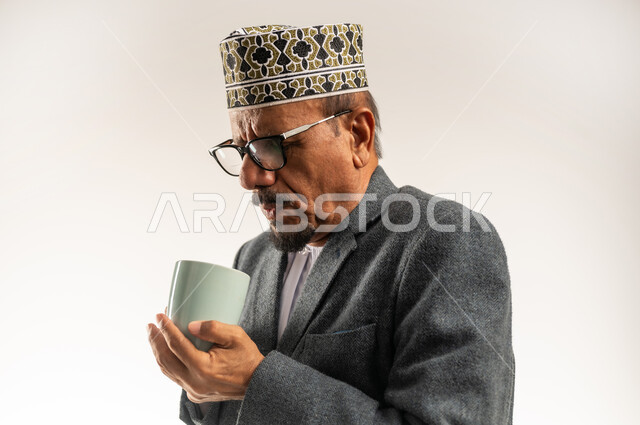 تناول كوب من القهوة الدافئة بالأجواء الشتوية الباردة، رفع حرارة الجسم وتدفئته، التغلب على الشعور بالبرد والصقيع، ارتداء ملابس الشتاء الثقيلة، بورتريه مقرب لرجل عربي خليجي عماني يرتدي الكمه والمعطف الشتوي يمسك فنجان المشروب الساخن، خلفية بيضاء
