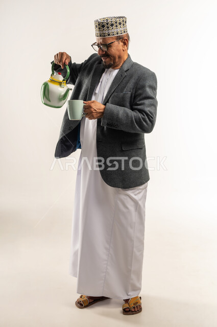 الاستمتاع بتناول مشروب ساخن، مقاومة الشعور بالبرد، رفع حرارة الجسم وتدفئته، الصقيع والأجواء الشتوية الباردة، ارتداء ملابس الشتاء الثقيلة، بورتريه لرجل عربي خليجي عماني يرتدي الكمه والمعطف الشتوي يقوم بصب الشاي من الابريق، الجسم بالطول الكامل، خلفية بيضاء