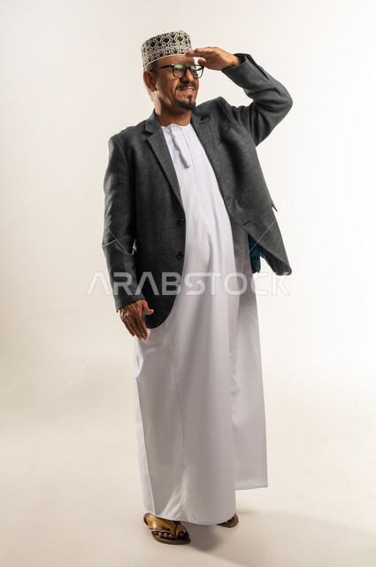 الرؤية الضبابية غير الواضحة، محاولة مشاهدة شيء ما بعيد، ارتداء ملابس الشتاء الثقيلة، الأجواء الشتوية القاسية، بورتريه لرجل عربي خليجي عماني يرتدي الكمه والمعطف الشتوي يقوم بوضع يده على جبينه، الجسم بالطول الكامل، خلفية بيضاء