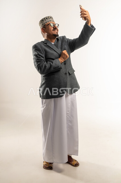 التقاط صورة سيلفي تذكارية, استخدام التقنيات التكنولوجية الحديثة والمتطورة, إيماءات البرودة والصقيع، بورتريه من الجانب لرجل عربي خليجي عماني يرتدي الدشداشه و الكمه والمعطف الشتوي يقوم بأخذ لقطة شخصية عبر الهاتف المحمول, الجسم بالطول الكامل, خلفية بيضاء