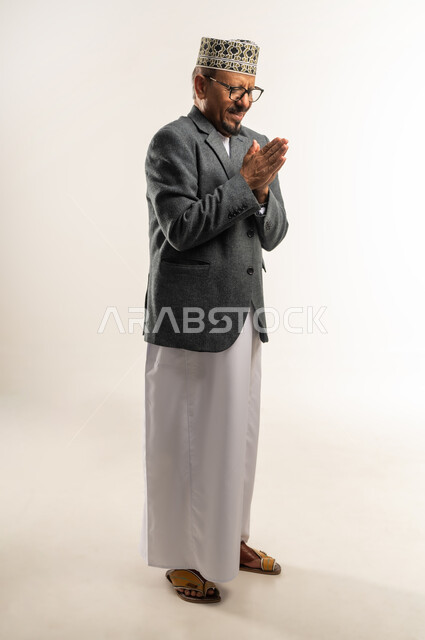 الارتجاف والشعور بالبرد، ارتداء الملابس الثقيلة لرفع حرارة الجسم، الأجواء الشتوية الباردة والصقيع، بورتريه من الجانب لرجل عربي خليجي عماني يرتدي الدشداشة والكمه والمعطف الشتوي يفرك كفيه ببعضهما لتدفئتهما، الجسم بالطول الكامل، خلفية بيضاء