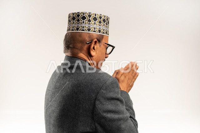 الصقيع والأجواء الشتوية الباردة، الارتجاف والشعور بالبرد، ارتداء الملابس الثقيلة لرفع حرارة الجسم، بورتريه مقرب من الخلف لرجل عربي خليجي عماني يرتدي الكمه والمعطف الشتوي يفرك كفيه ببعضهما لتدفئتهما، خلفية بيضاء