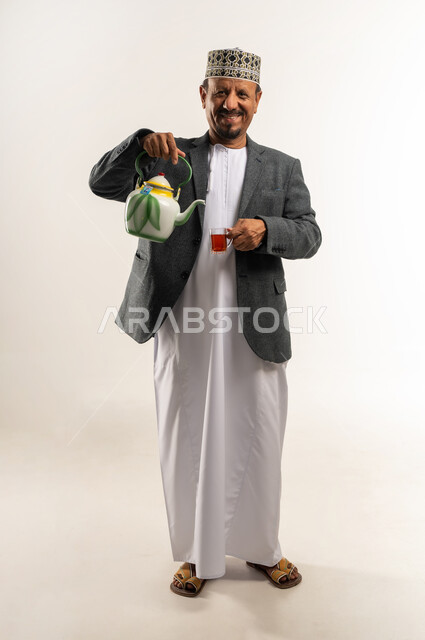 مواجهة البرد بارتداء ملابس الشتاء الثقيلة، تدفئة الجسم ورفع حرارته عن طريق احتساء مشروب ساخن، الصقيع والأجواء الشتوية الباردة، بورتريه لرجل عربي خليجي عماني يرتدي الكمه والمعطف الشتوي يقوم بصب الشاي من الابريق بإيماءات السرور، الجسم بالطول الكامل، خلفية بيضاء