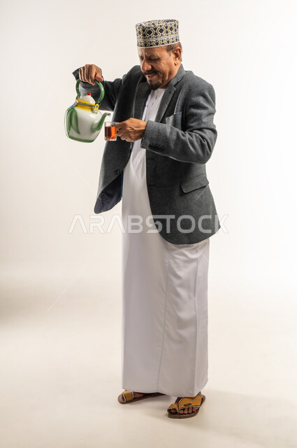 تدفئة الجسم ورفع حرارته عن طريق احتساء مشروب ساخن، الصقيع والأجواء الشتوية الباردة، ارتداء ملابس الشتاء الثقيلة، بورتريه لرجل عربي خليجي عماني يرتدي الكمه والمعطف الشتوي يقوم بصب الشاي من الابريق، الجسم بالطول الكامل، خلفية بيضاء